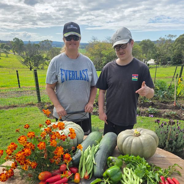 Two people standing behind a table of fresh vegetables and flowers outdoors.