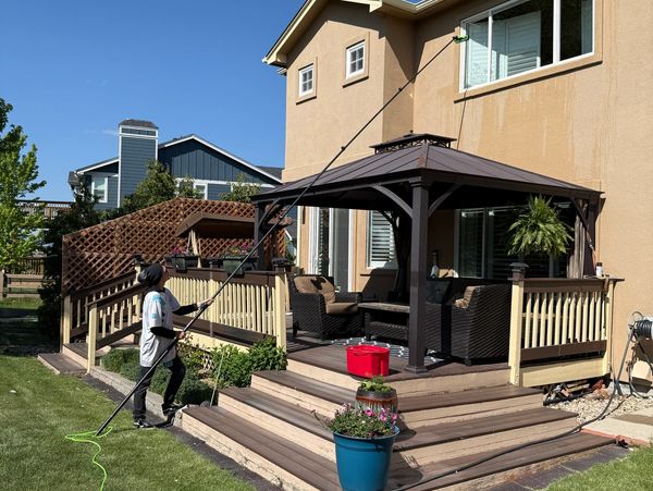 A person cleans a house window using an extended pole from the backyard deck.