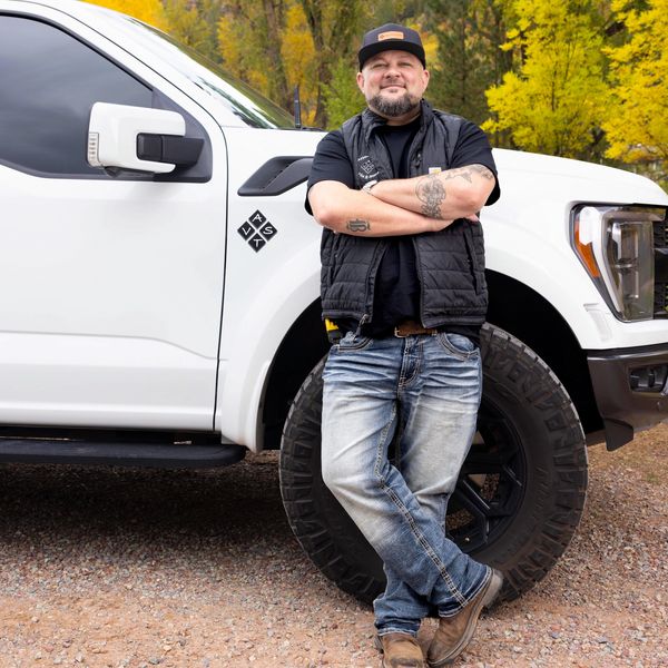 Man with tattoos leaning against a white truck in autumn scenery.