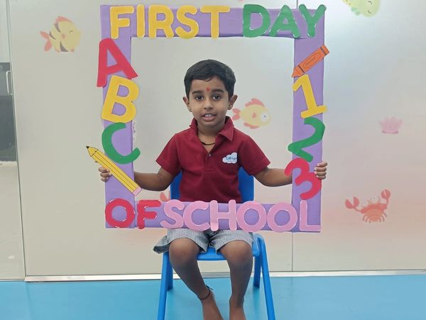Young boy sitting on a blue chair holding a colorful "First Day of School" frame.