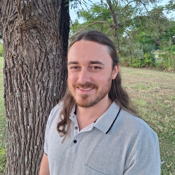 Smiling man with long hair and beard stands by a tree in a park.