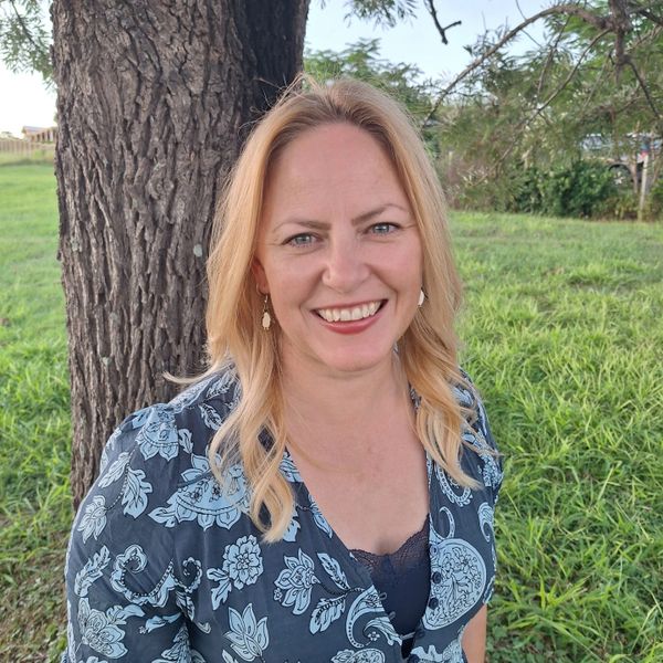 Smiling woman with blonde hair standing by a tree in a patterned dress.