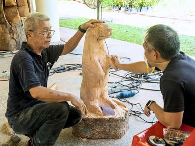 Two men sculpt a large wooden animal figure outdoors.