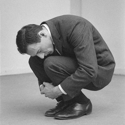 Man in a suit crouching tightly on the floor in a minimal room.