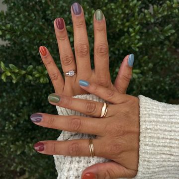 Hands with multicolored nail polish and rings against a leafy background.
