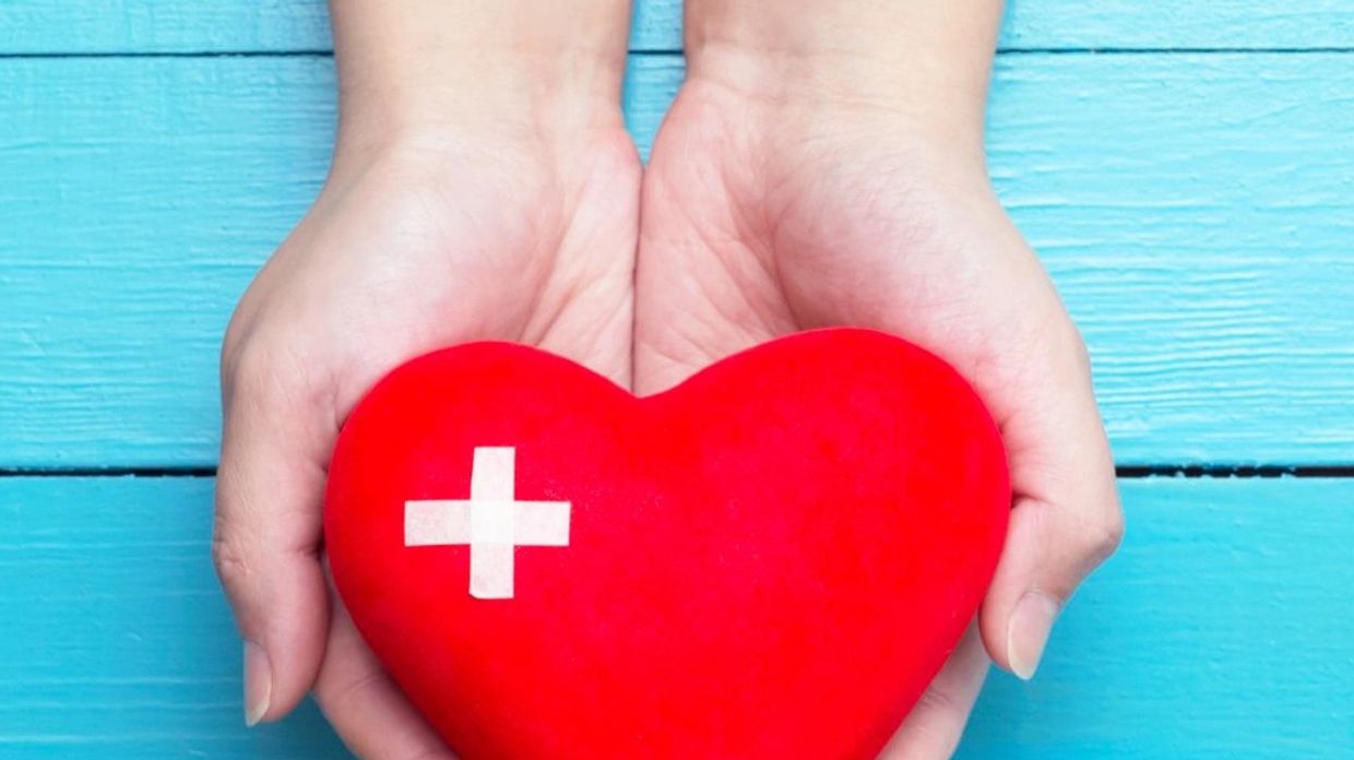 Hands holding a red heart with a white medical cross on a blue wooden background.