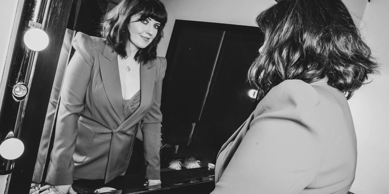 Woman with dark hair and blazer smiling at her reflection in a mirror with lights.