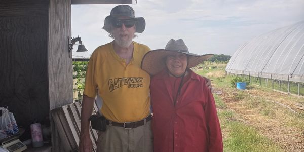 An elderly couple wearing hats stands outdoors near a greenhouse on a sunny day.