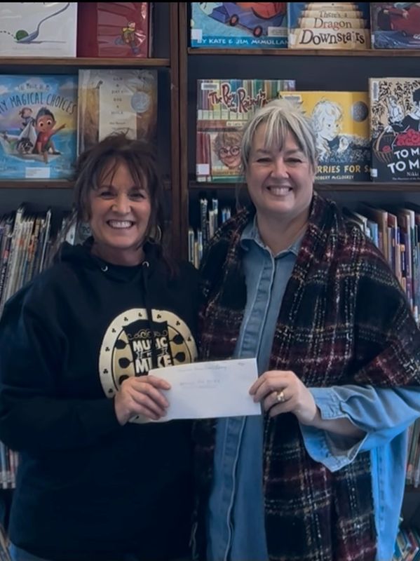 Two smiling women holding an envelope in front of a library bookshelf.