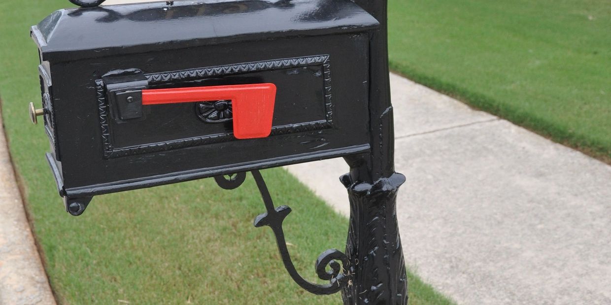 Black mailbox with a red flag on a decorative post near a sidewalk.