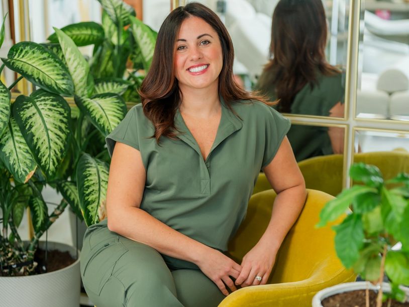 Woman in green outfit smiling while seated by plants and a yellow chair.