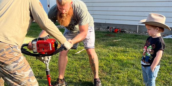 Two men using a red auger while a child in a cowboy hat watches.