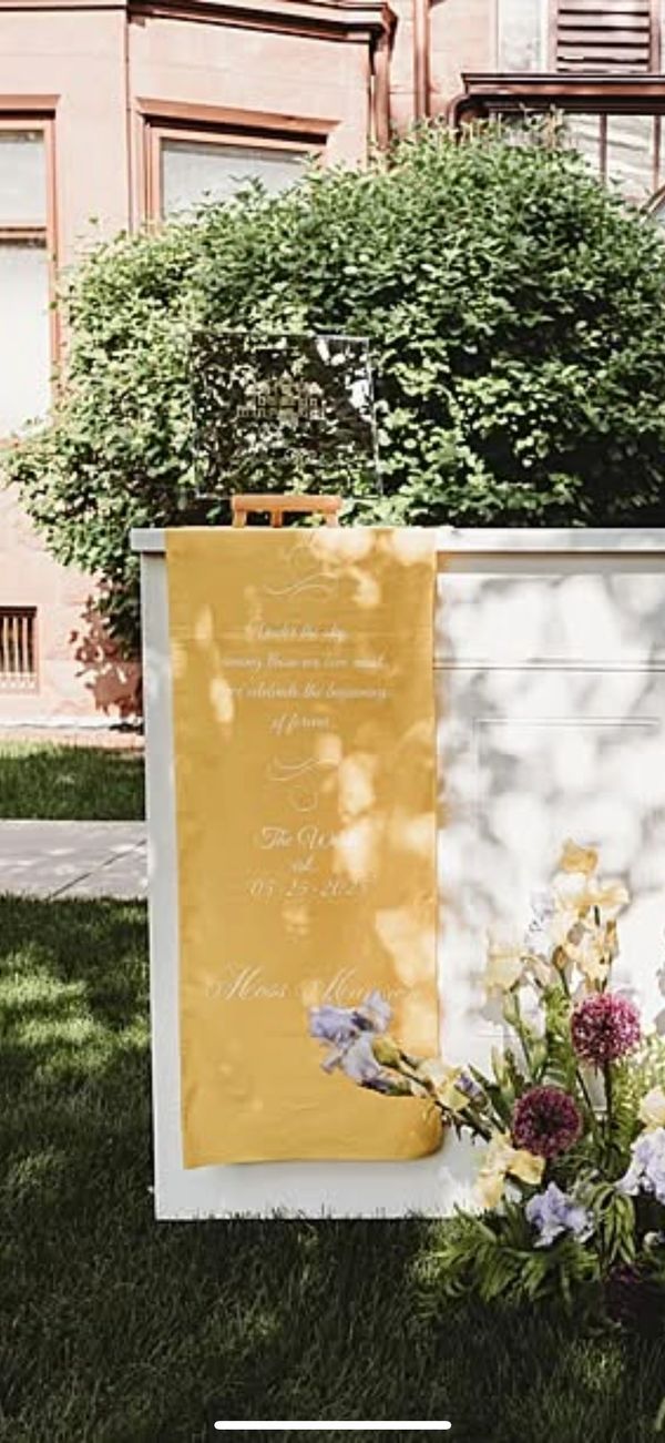 Outdoor wedding welcome sign with yellow fabric and floral arrangement.