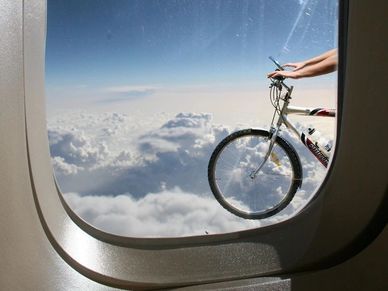 Person holding a bicycle outside an airplane window above the clouds.