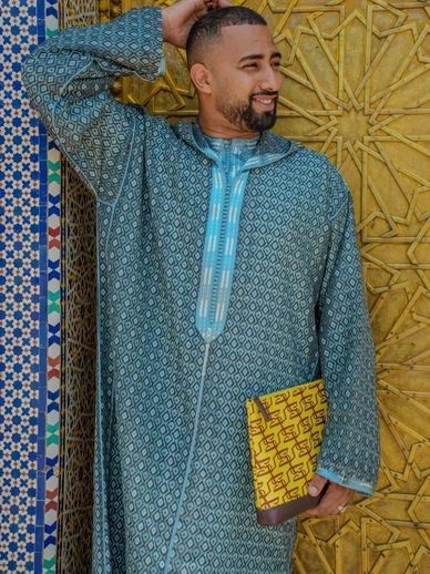 Man in traditional attire holding a patterned clutch smiling against an ornate wall.