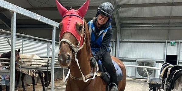 Smiling rider on a chestnut horse wearing a red mask in an indoor stable.