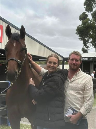 Two people smiling with a horse at a stable area on a cloudy day.