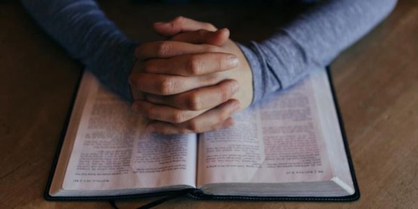 Person with clasped hands resting on an open book, possibly praying or reflecting.