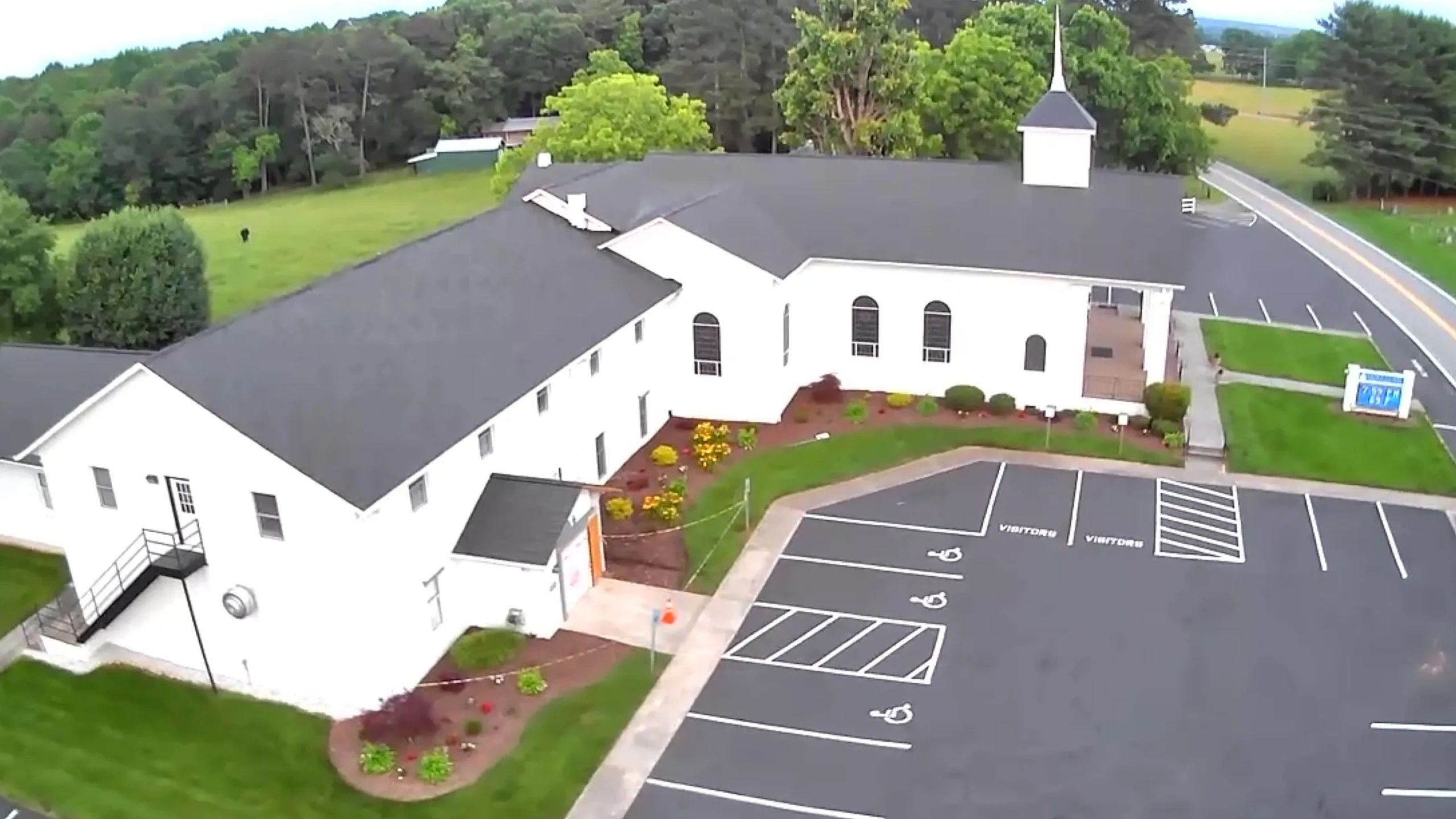A white church building with a black roof and empty parking lot on a green landscape.