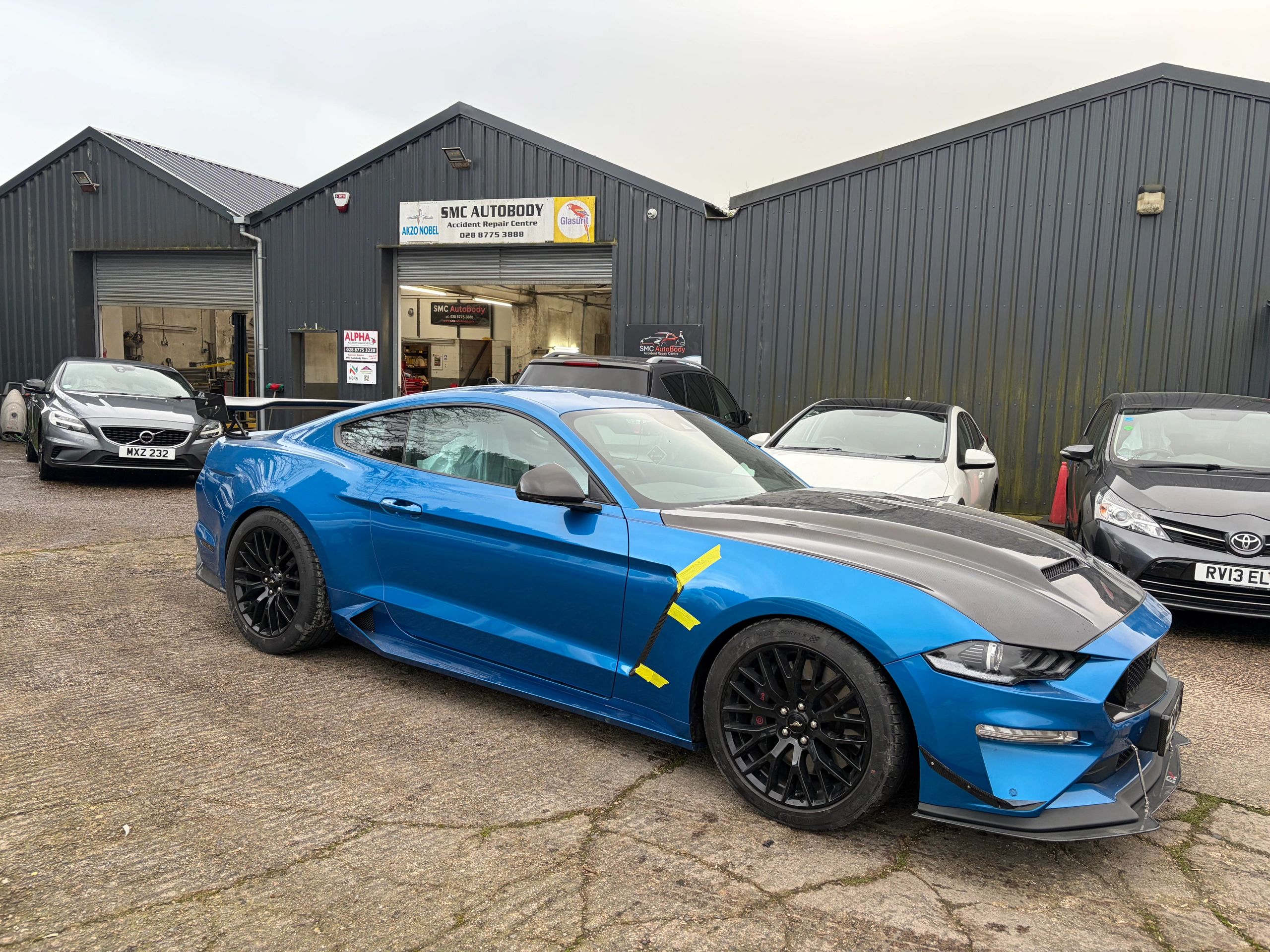 Blue sports car with black hood parked outside an auto repair shop.