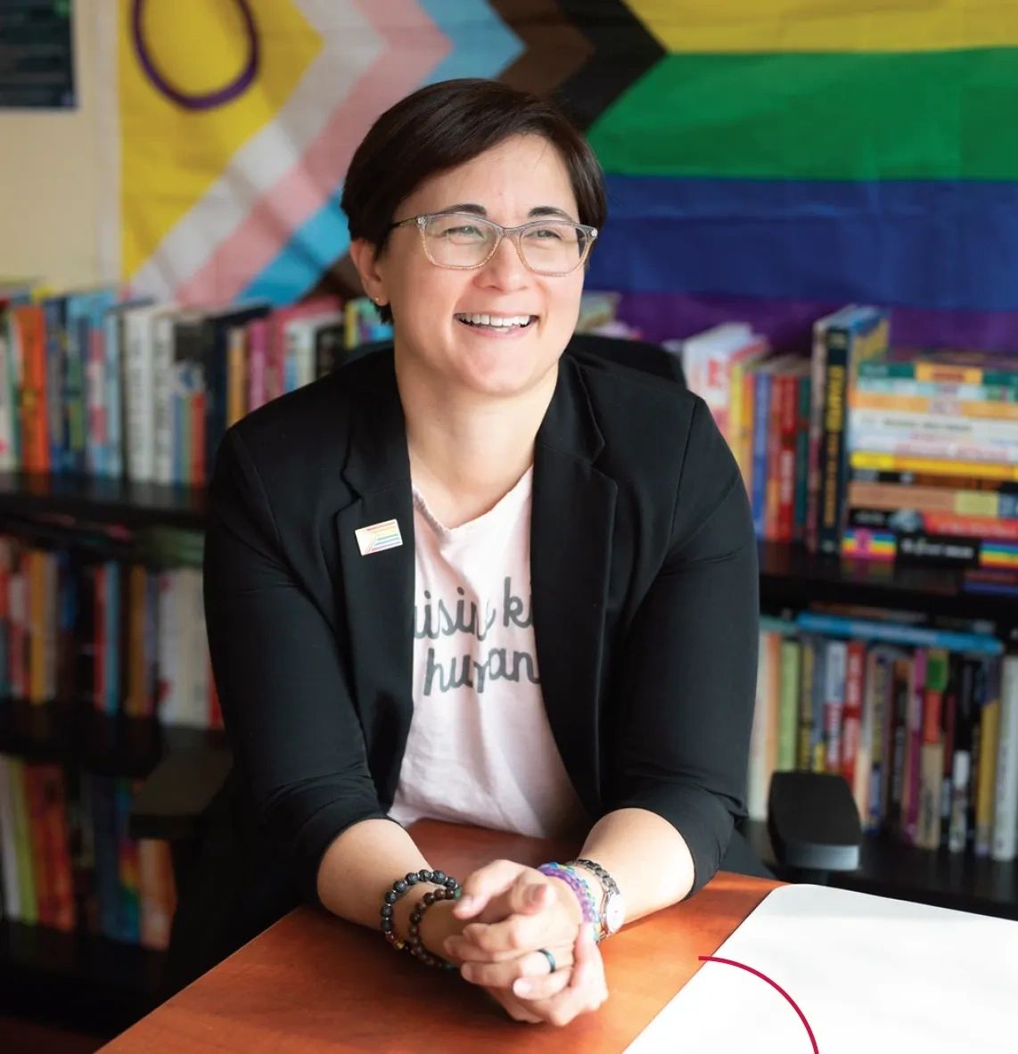 Smiling person with glasses sitting at a desk with books and a pride flag behind.