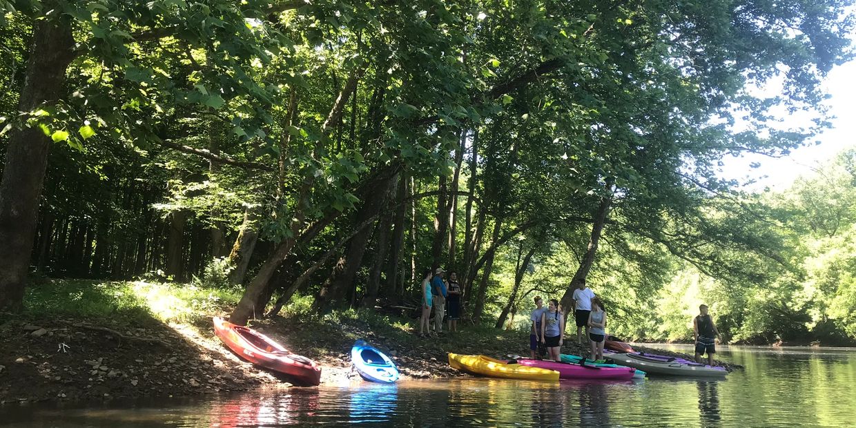 People preparing kayaks by a riverside surrounded by lush green trees.