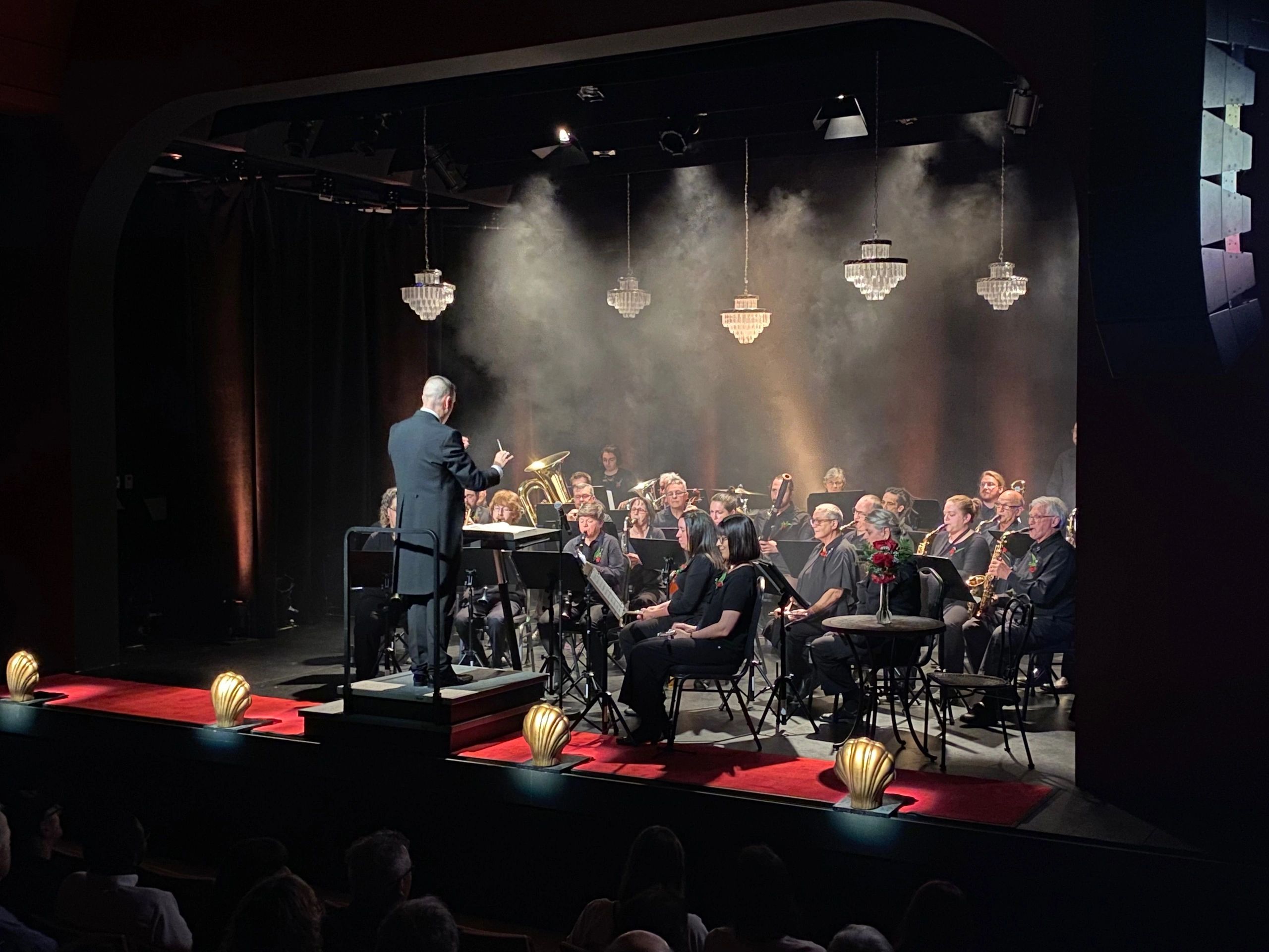 Conductor leads an orchestra on a dimly lit stage with chandeliers and smoke effects.