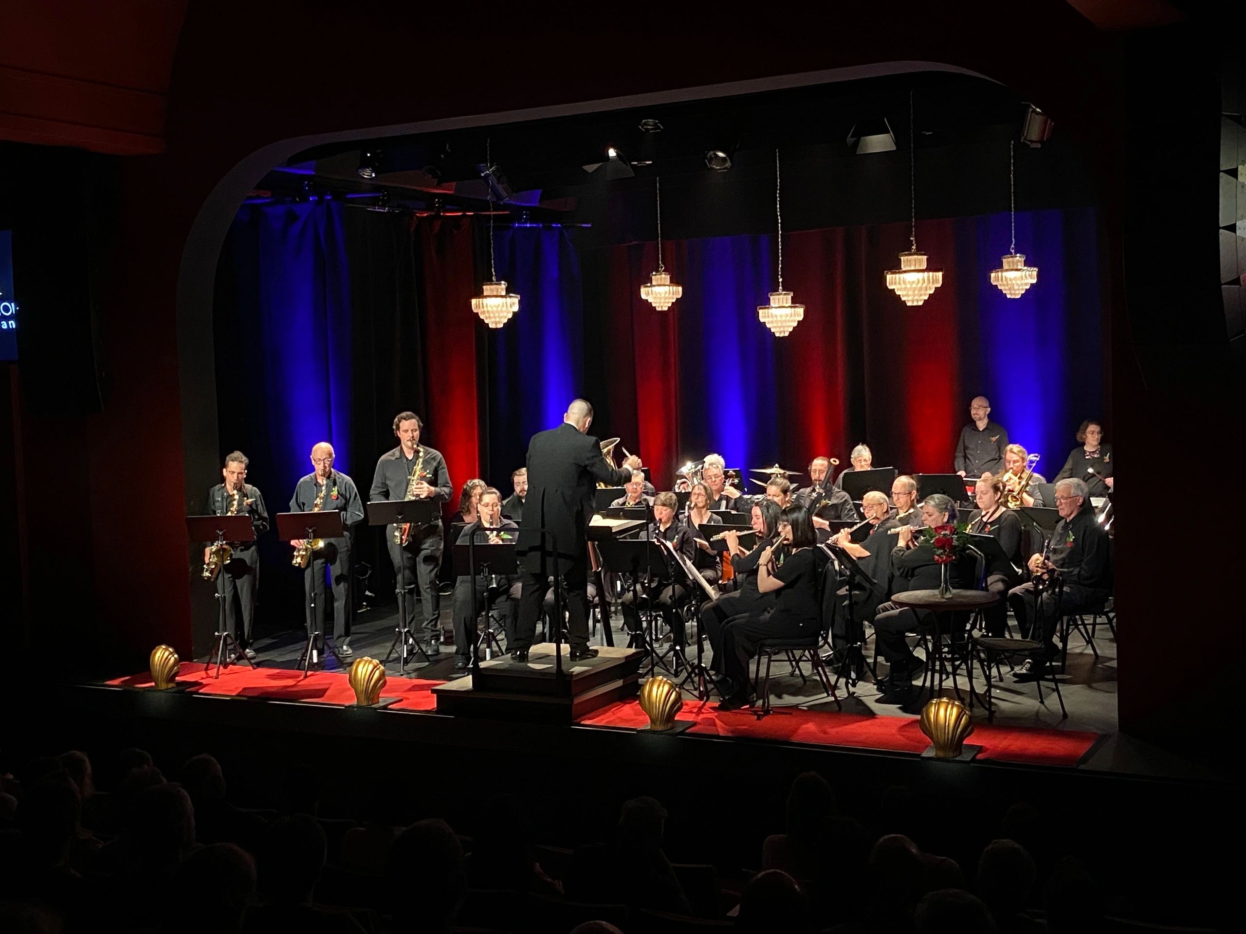 Concert band performing on stage with colorful lighting and chandeliers.