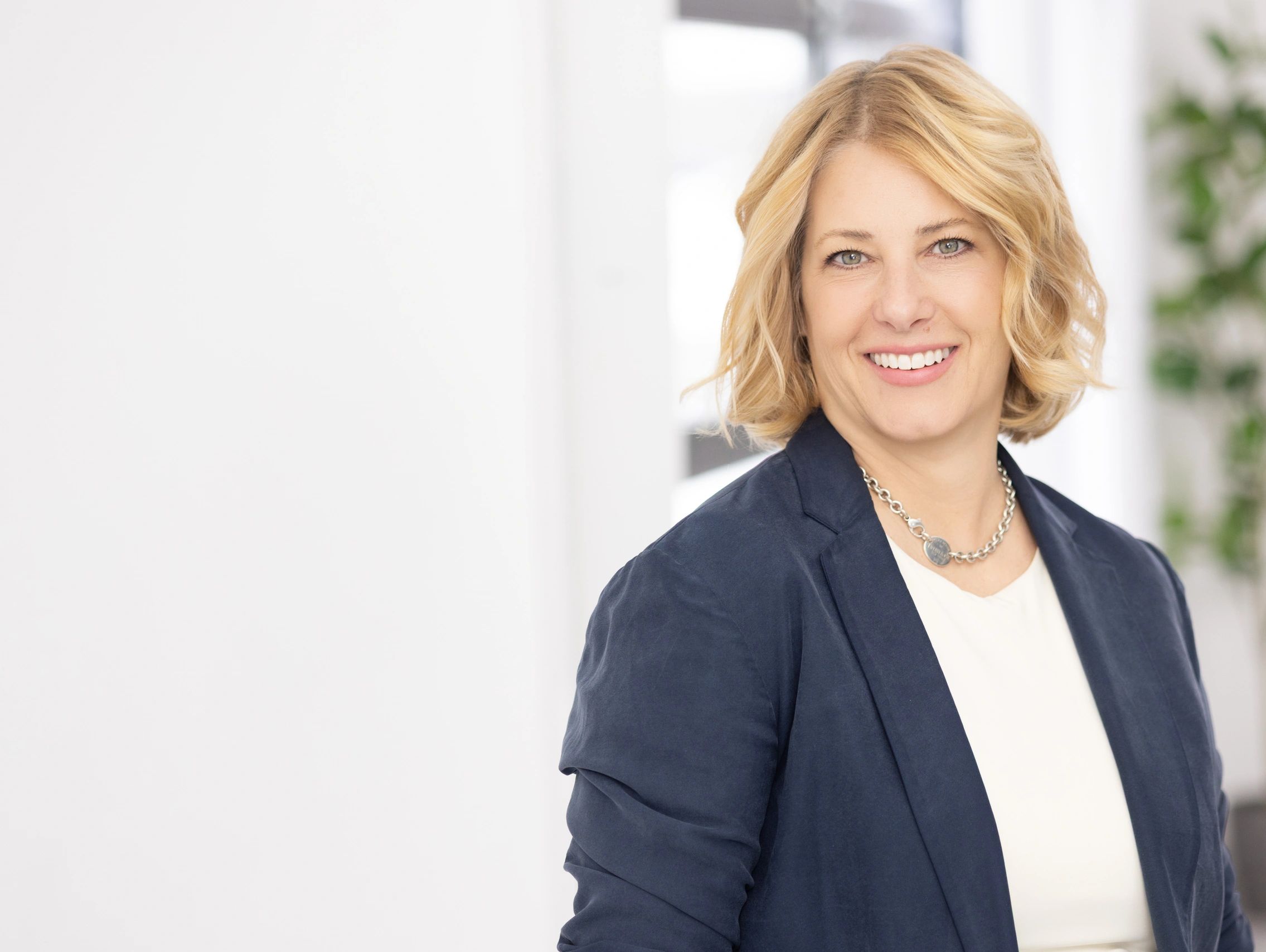 Smiling woman with blonde hair in a navy blazer and chain necklace.