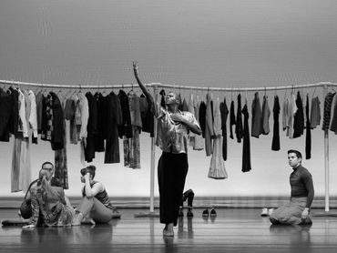 A black and white image of dancers in a theatrical performance with clothes hanging in the background.