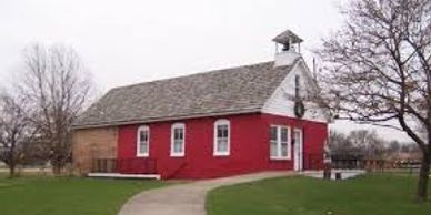 A small red and white schoolhouse with a bell tower in a grassy area.