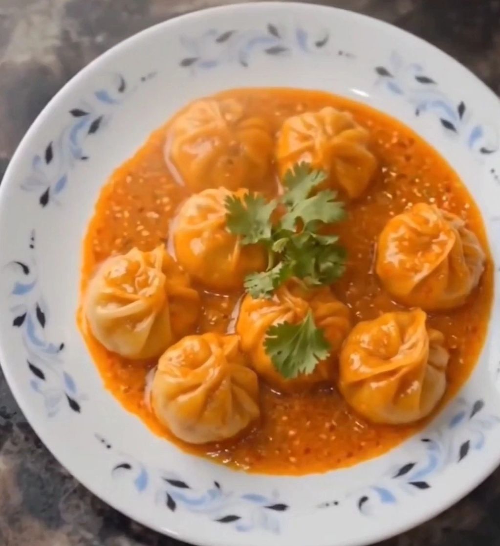 Plate of steamed momos in spicy orange sauce, garnished with cilantro.