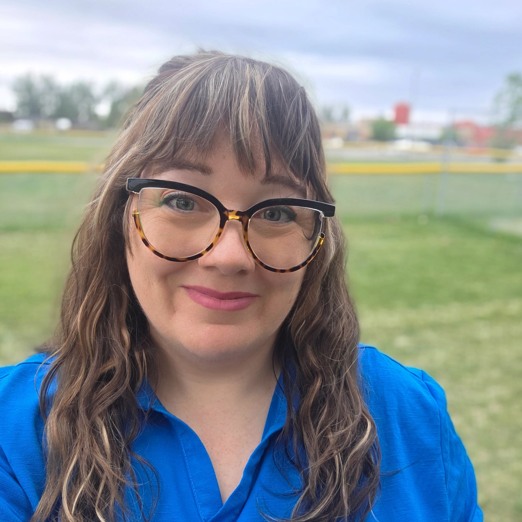 Smiling woman with glasses and wavy hair in a blue shirt outdoors.