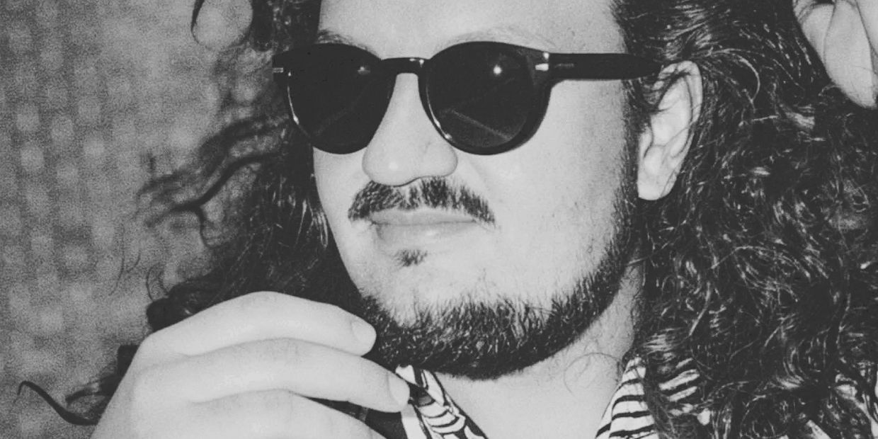 Man wearing sunglasses with long curly hair and facial hair.