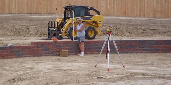 Construction worker with surveying equipment near a yellow loader and brick wall.