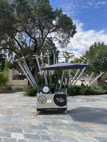 Mobile coffee cart under metal sculpture on a sunny day.