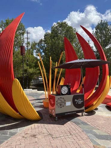 Mobile coffee cart under a black umbrella near colorful outdoor sculpture.