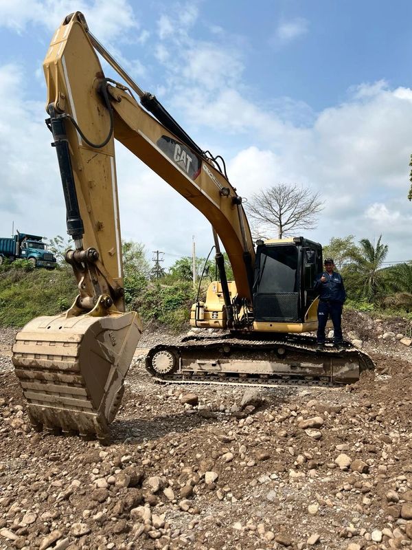 Large CAT excavator on rocky ground with worker standing beside it.