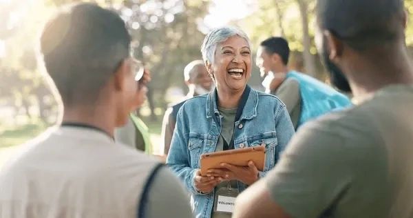 A joyful senior volunteer laughing and holding a tablet outdoors with a group.