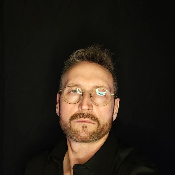 Man with glasses and beard against black background.