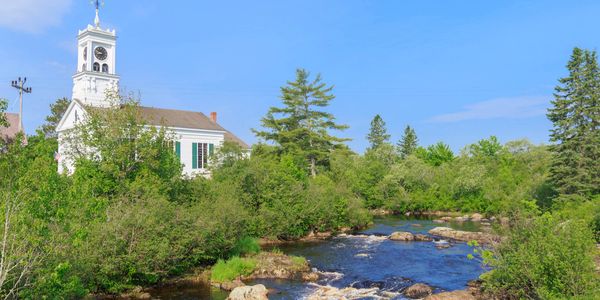 A white church with a clock tower by a river surrounded by greenery under a blue sky.