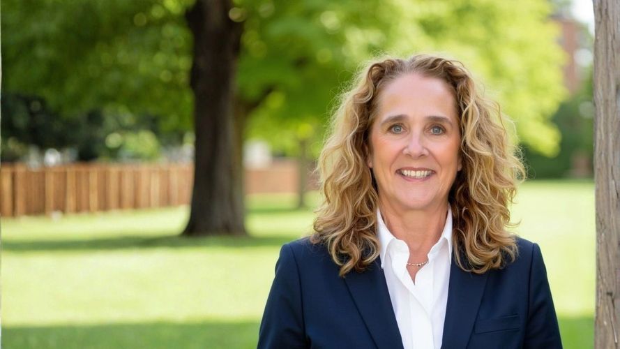 Smiling woman in a navy blazer stands outdoors with trees and grass behind her.