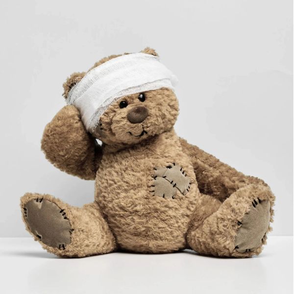 A teddy bear with a bandaged head and stitched patches sitting on a white surface.