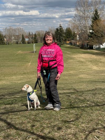 A woman in a pink Saskatoon hoodie stands smiling with her dog in a park.
