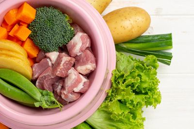 Fresh ingredients including meat, vegetables, and eggs arranged in and around a pink bowl.