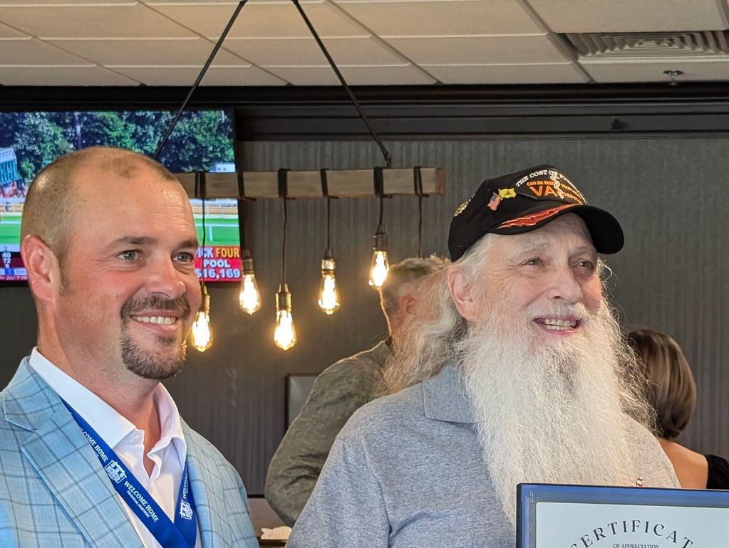 Two men smiling, one wearing a veteran cap and holding a certificate.