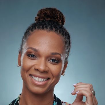 Smiling woman with braided hair in a bun, wearing pearl earrings and a patterned top.