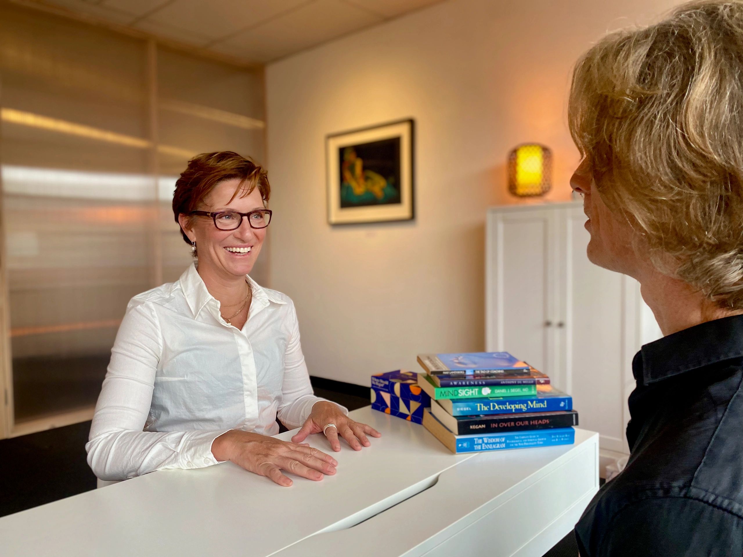 Two women conversing in an office setting, books on the desk.