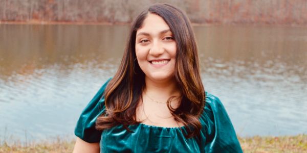 A smiling woman in a teal dress stands by a lake.