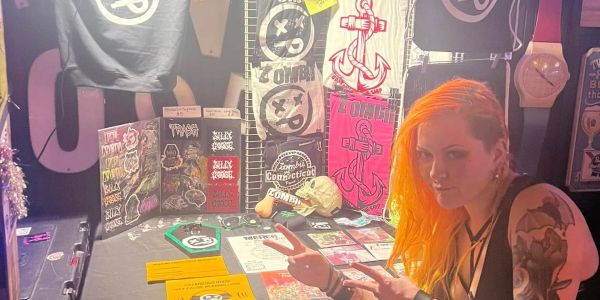 Person with orange hair and tattoos at a merch booth with punk-themed items.